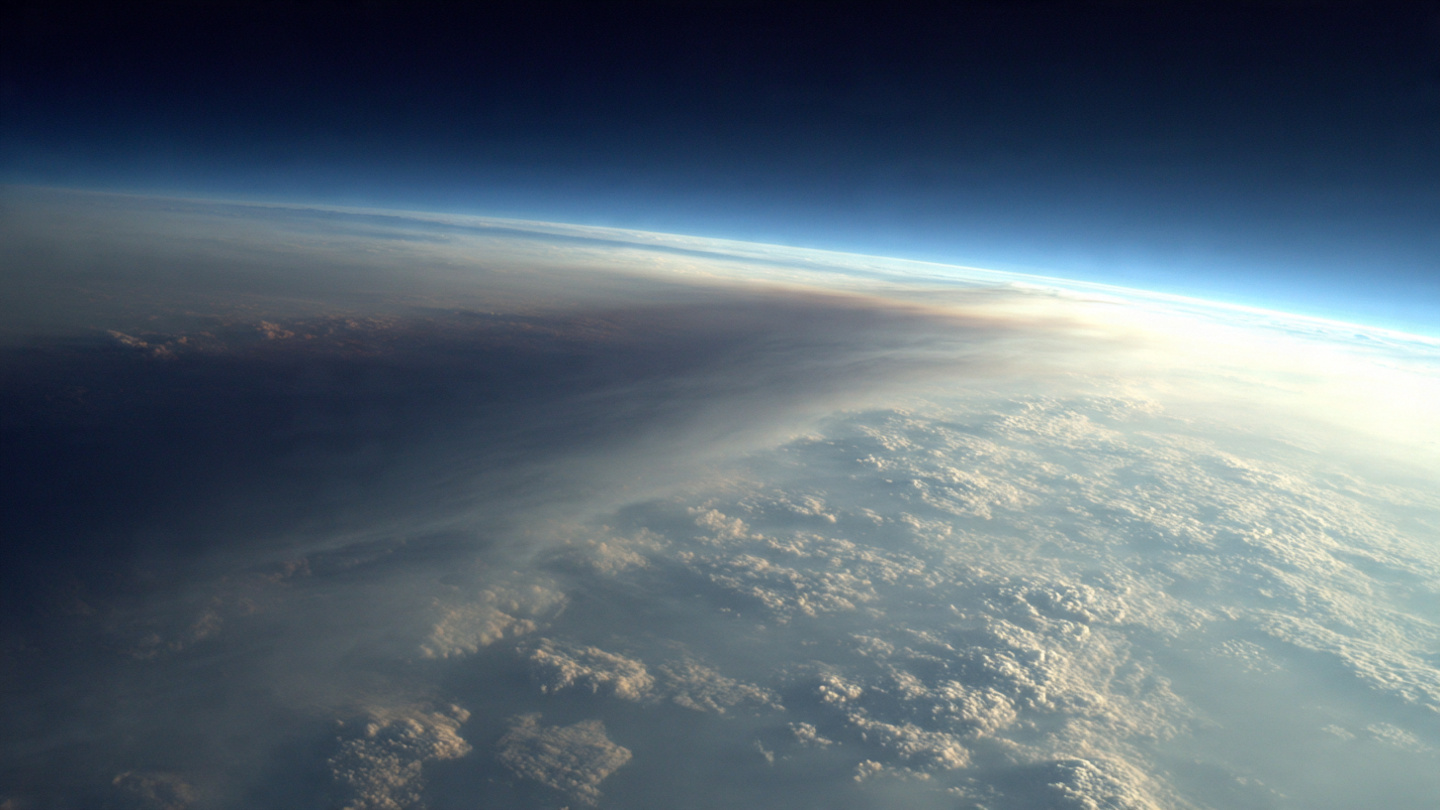 Aerial view of stratospheric particles dispersing above Earth's atmosphere, sun partially obscured by a thin reflective haze layer
